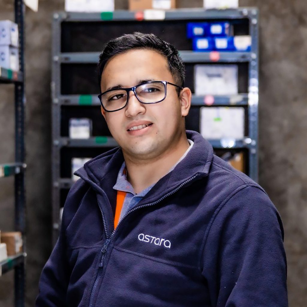 Hombre joven con gafas y chaqueta azul con el logo de Astara, en un entorno de bodega con estanterías metálicas y cajas organizadas detrás de él.