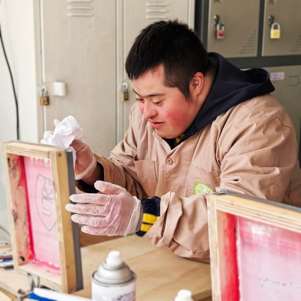Hombre joven con síndrome de Down trabaja concentrado en un taller de serigrafía, usando guantes de látex y manipulando un marco de impresión sobre una mesa con materiales.