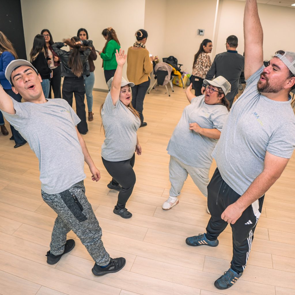 Cuatro personas con discapacidad intelectual bailan y levantan los brazos con entusiasmo en una sala, mientras al fondo otras personas observan y conversan.