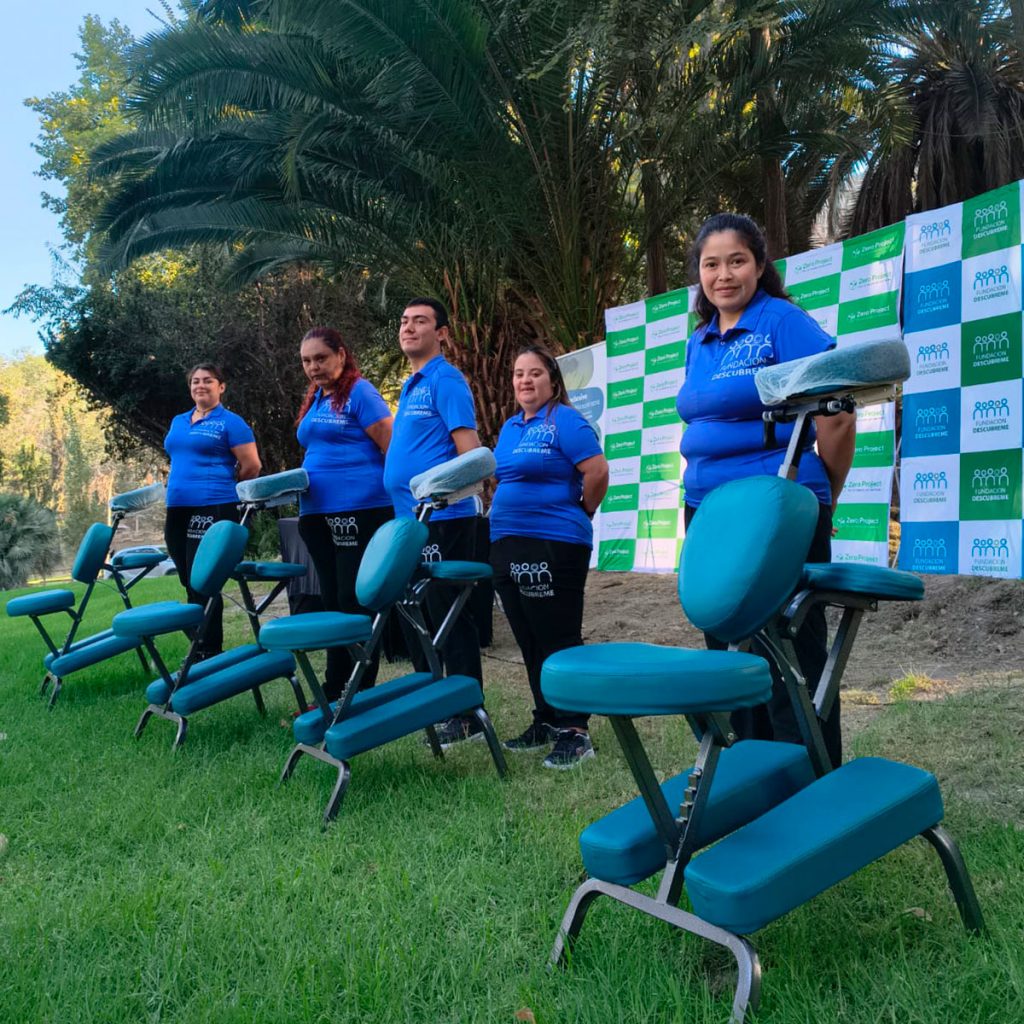 Equipo de masoterapia inclusiva de Fundación Descúbreme en una actividad al aire libre. Cinco personas con polera azul institucional posan detrás de varias sillas de masaje sobre el pasto, con palmeras y un panel de fondo.